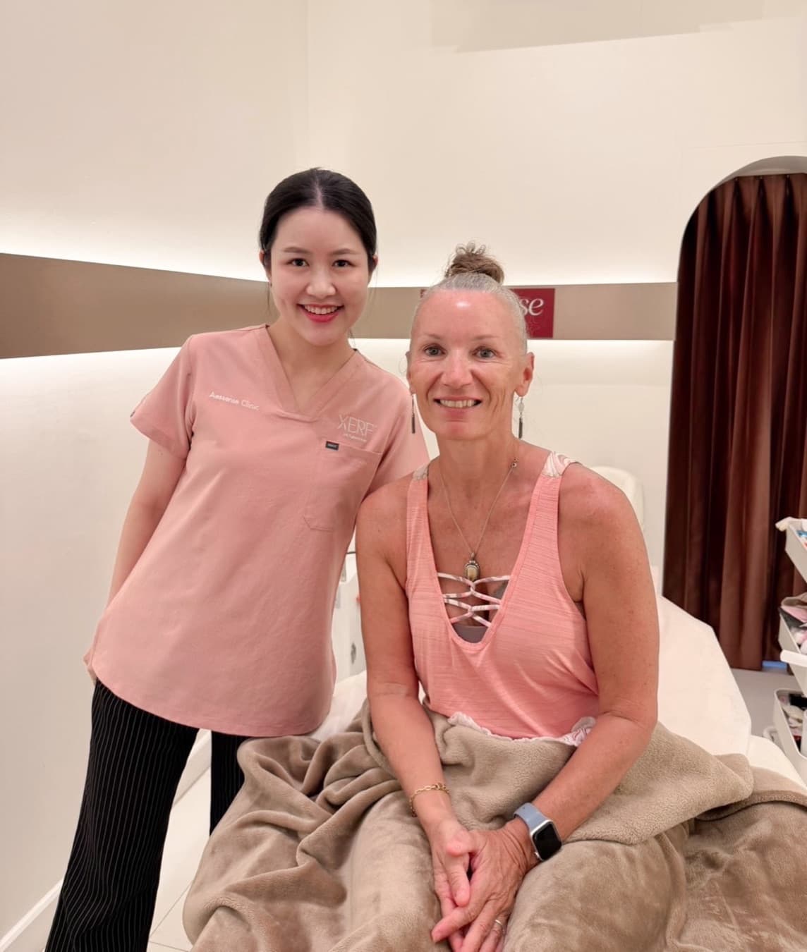 A smiling woman in pink AESsense scrubs stands beside a smiling client seated on a treatment bed with a blanket over her lap